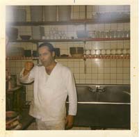 Nikolas dressed as a white cook, standing in front of a sink and kitchen utensils.