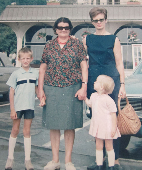 An older woman holds the hands of a small boy and girl, while another woman stands beside her.
