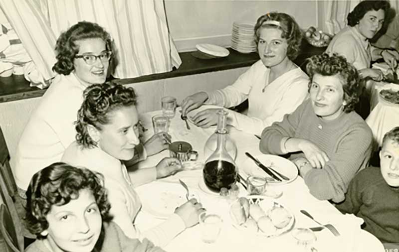 Four women and two children are seated around a dinner table.