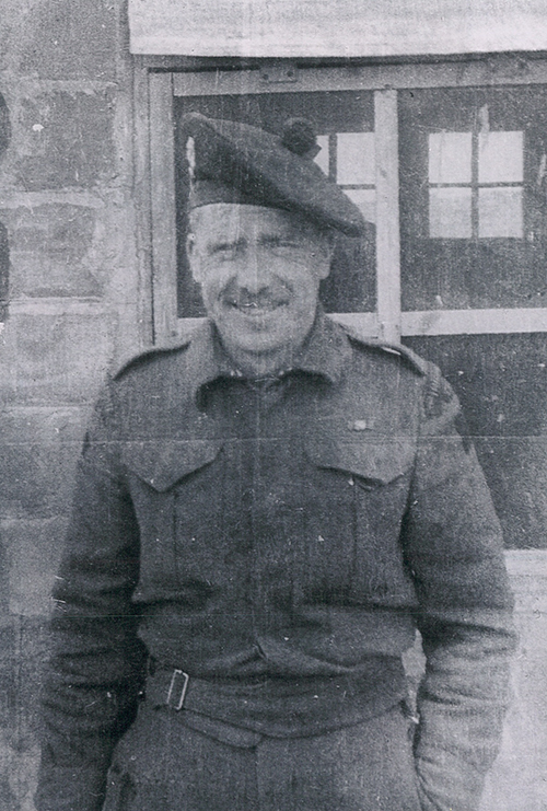 A man stands in front of a building, he is dressed in military clothing.