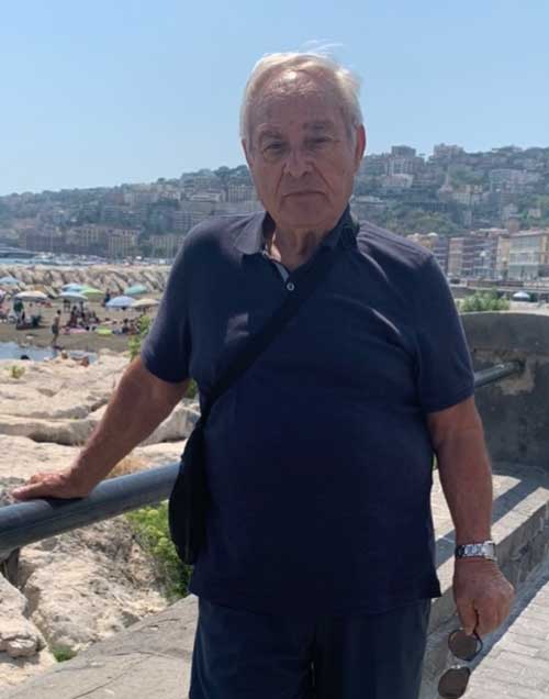 An older man in a blue t shirt stands against a railing, there is a scenic background behind him.