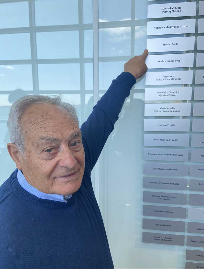 A man points to a plaque on a wall with his name on it.