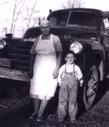 Woman in apron and a little boy in overalls leaning against an old car.