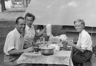 Black and white photo of three people eating food while camping.