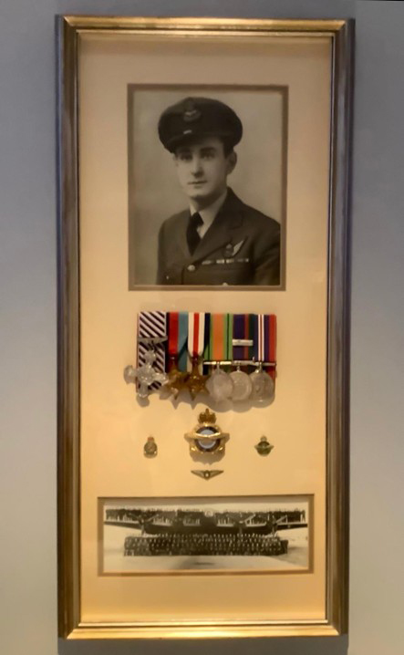 A color photograph of a picture and memorabilia in a frame hanging on the wall, the frame contains Les Gallant's official photograph in his uniform, his service medals, and a black-and-white photograph of his RAF unit in front of a bomber.